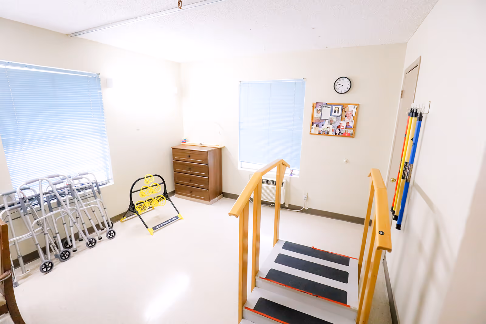 A bright rehabilitation room with two windows covered by blue blinds, several walkers lined up against the wall, a wooden chest of drawers, a bulletin board with photos and notes, a clock on the wall, and a small set of stairs with handrails for physical therapy exercises. Resistance bands are hanging on the wall near a closed door.
