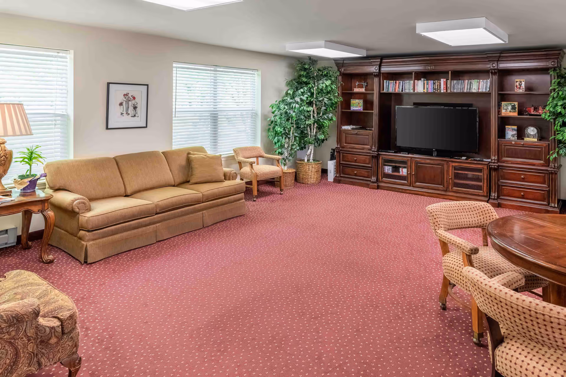 A cozy living room with a brown sofa, two armchairs, a wooden entertainment center with a flat-screen TV, books, and decorative items. The room has two large windows with white blinds, a side table with a lamp and plants, and a red carpet with a subtle pattern. There are also two large potted plants in the corners.