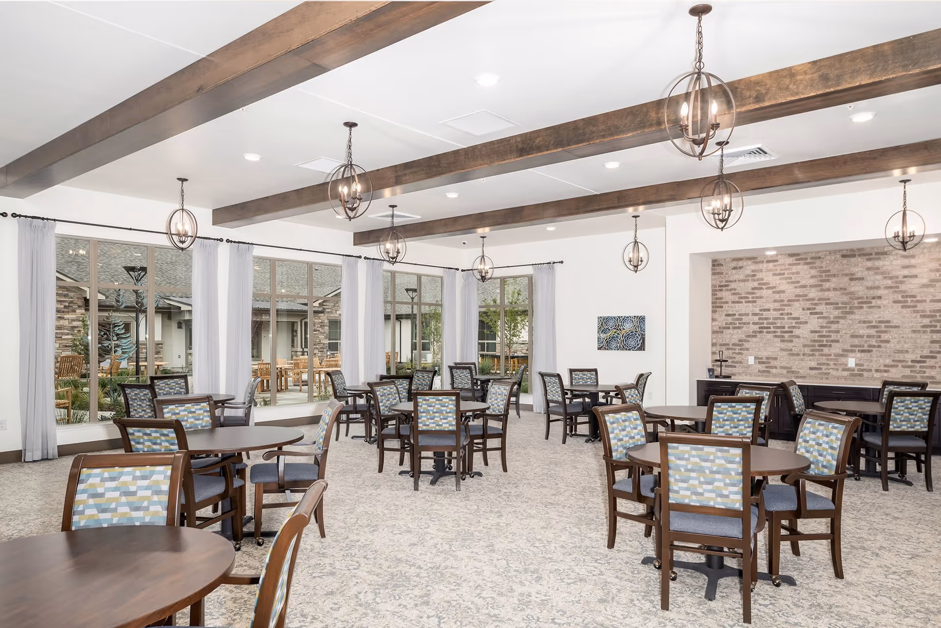 A bright and spacious dining room in a senior living facility with multiple round wooden tables and cushioned chairs featuring patterned backs. The room has large windows with sheer white curtains, wooden ceiling beams, and modern spherical pendant lights. Outside the windows, there is a view of an outdoor patio area with wooden benches and greenery.