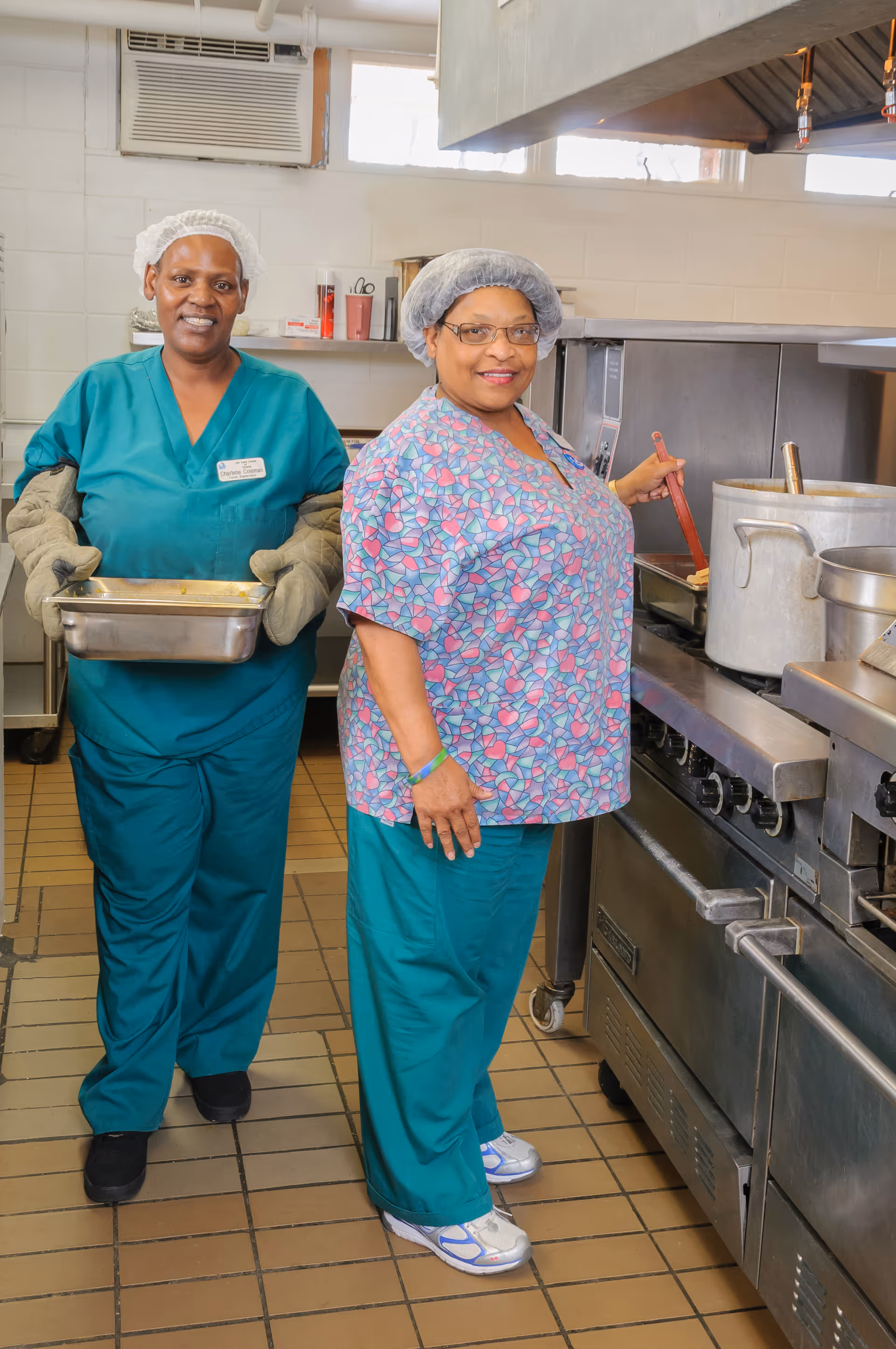 Two women in a commercial kitchen. One woman is stirring food in a large pot on the stove while wearing a hairnet, glasses, a patterned scrub top, and teal pants. The other woman is holding a metal tray with oven mitts, wearing a hairnet and teal scrubs. The kitchen has stainless steel appliances and a tiled floor.