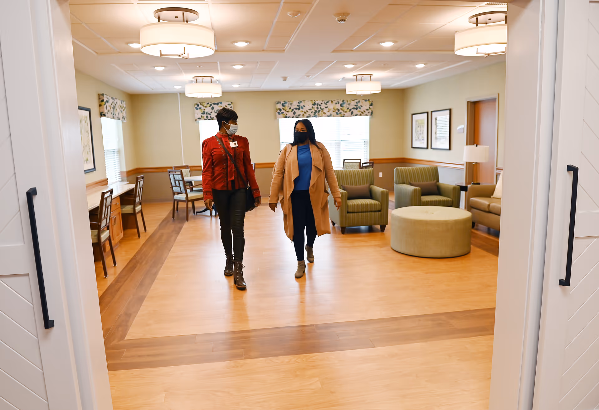 Two women wearing face masks walking through a well-lit common area with wooden floors, green armchairs, a round ottoman, and tables with chairs near the windows.