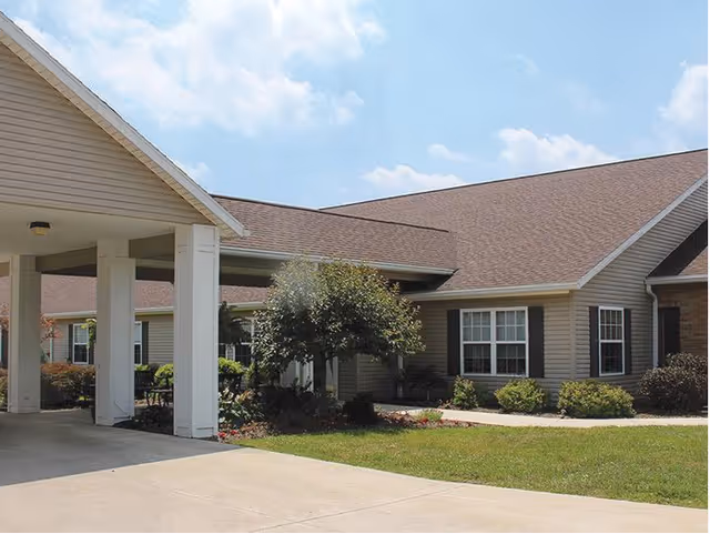 Exterior view of a single-story building with beige siding and a brown roof under a partly cloudy sky. The building has multiple windows with white frames and black shutters. There is a covered driveway entrance supported by white columns, a small tree, and landscaped bushes in front of the building.