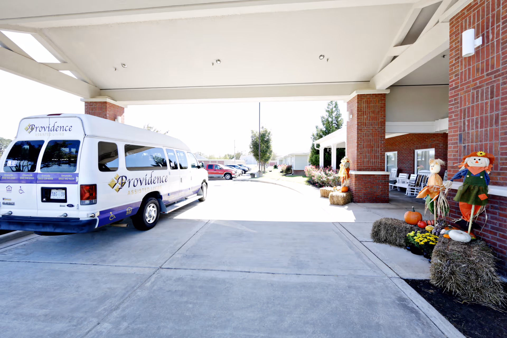 Covered front entrance of Providence Assisted Living with the facility shuttle van parked and fall decorations on the porch.