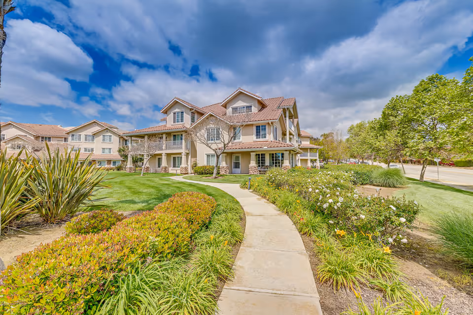 A bright and sunny exterior view of The Orchard Senior Living facility showing a multi-story building with beige walls and a tiled roof. The foreground features a curved concrete walkway bordered by well-maintained green lawns, bushes, and flowering plants. Trees line the right side of the walkway under a partly cloudy blue sky.