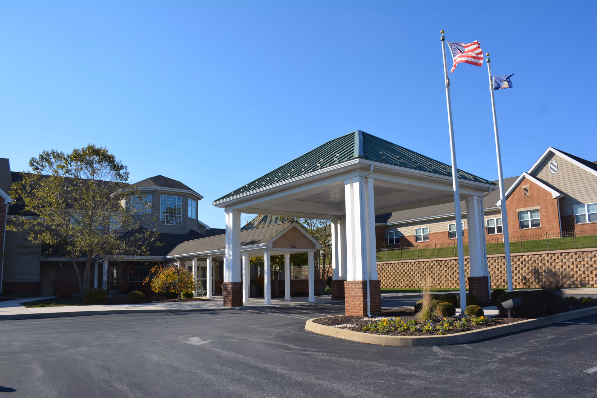 Front porte-cochère and entrance of a senior living building with flagpoles, landscaping, and brick-and-siding wings under a clear blue sky.
