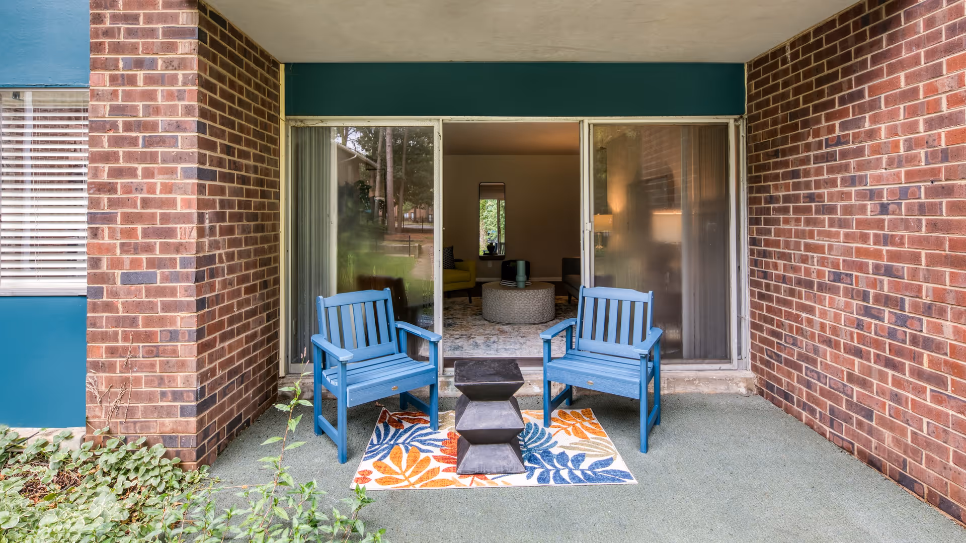 A small outdoor patio area with two blue wooden chairs and a black geometric side table placed on a colorful leaf-patterned rug. The patio is surrounded by brick walls and has a sliding glass door leading into a living room with a yellow chair, a round ottoman, and a tall mirror on the wall.