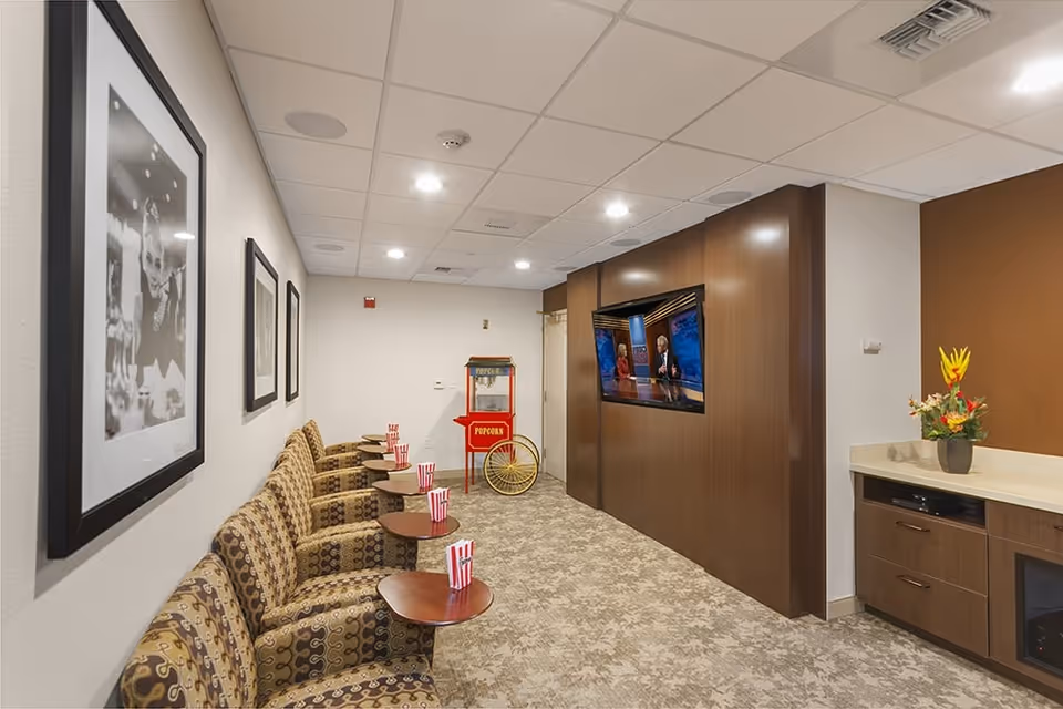 Cozy media/lounge room with patterned armchairs and small round tables facing a wall-mounted TV and a popcorn cart.