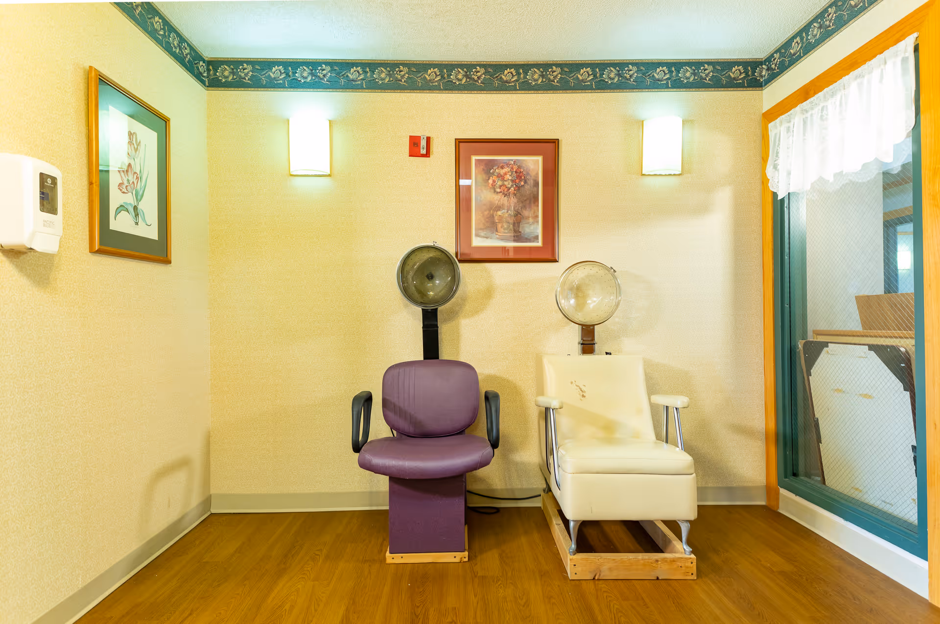 Two hooded hair-dryer salon chairs (one purple, one beige) in a small interior room with framed artwork and wall sconces.