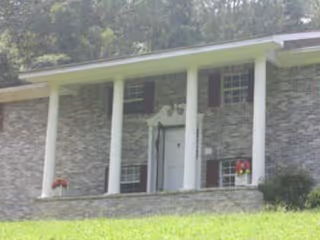 Front exterior view of a single-story building with gray brick walls, white columns, and a white door with decorative trim. There are windows with dark shutters on either side of the door and flower pots with red flowers on the porch.