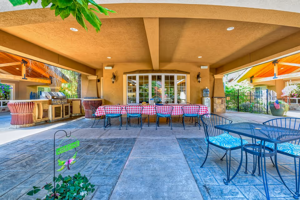 Outdoor covered patio area with a long table covered in a red and white checkered tablecloth surrounded by chairs. To the left, there is a built-in grill and countertop area. To the right, there is a metal table with blue patterned cushions on the chairs. The area is paved with stone tiles and there are plants and greenery visible around the patio.