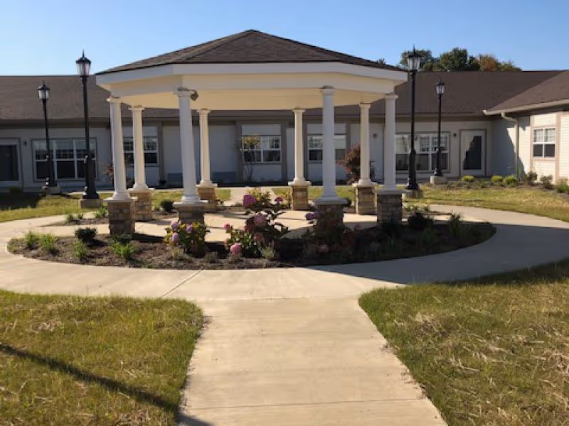 Outdoor courtyard area of a senior living facility featuring a circular paved walkway surrounding a garden bed with flowers and a gazebo supported by white columns. The building with multiple windows and a brown roof surrounds the courtyard.