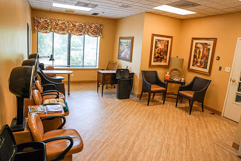 A bright senior living common room with chairs and small tables arranged around a window and framed artwork on beige walls.