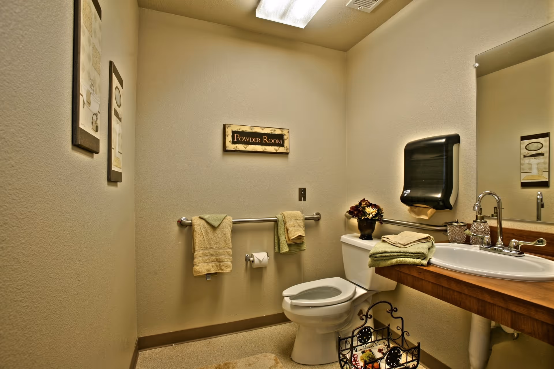 A small, clean powder room with a toilet, a wooden countertop with a sink, a wall-mounted paper towel dispenser, and a mirror. There are towels hanging on a grab bar next to the toilet and folded towels on the countertop. The walls are decorated with framed pictures and a sign that reads 'Powder Room'.