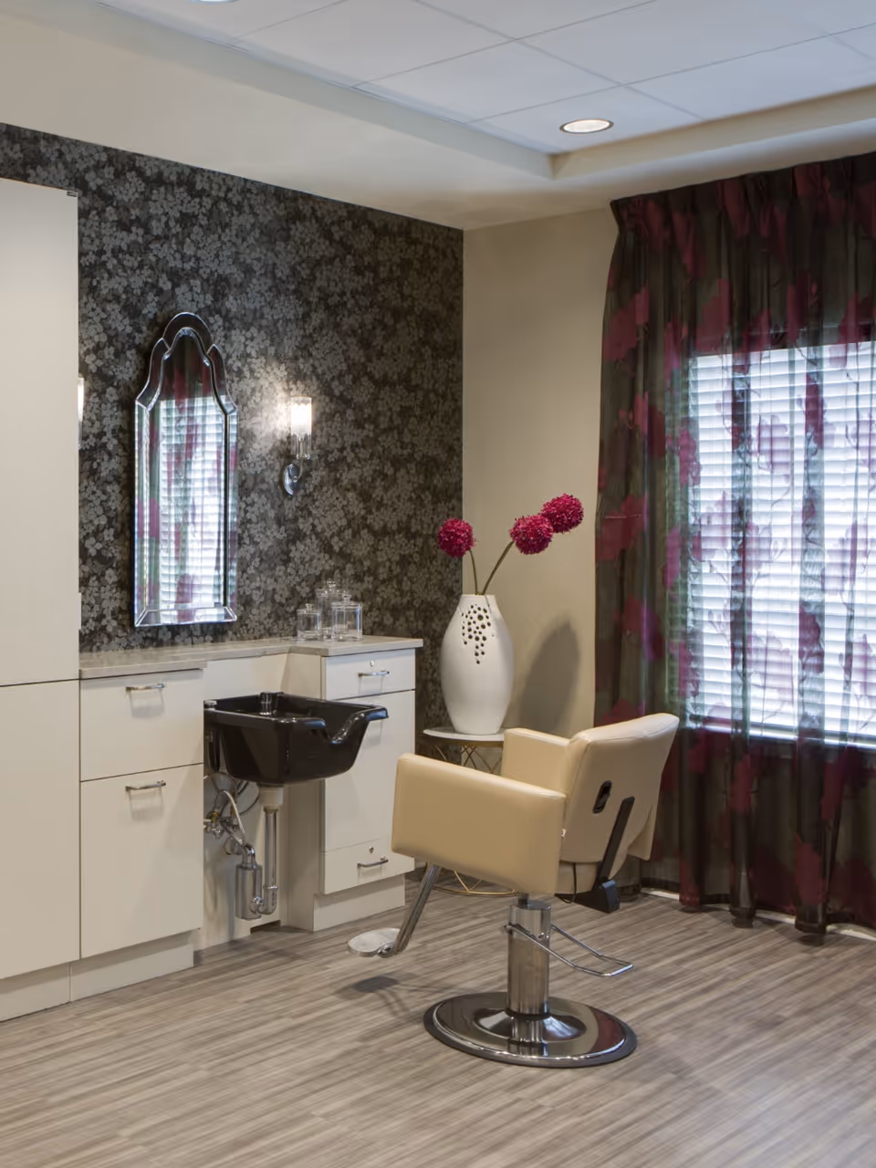 A salon styling station with a beige swivel chair, black wash basin, mirrored wall, cabinets and a vase of purple flowers by a curtained window.