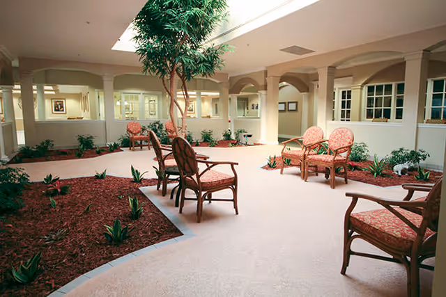 Indoor courtyard area with a large skylight, several wooden chairs with red patterned cushions arranged around the space, small plants in mulched garden beds, and a tall tree in the center.