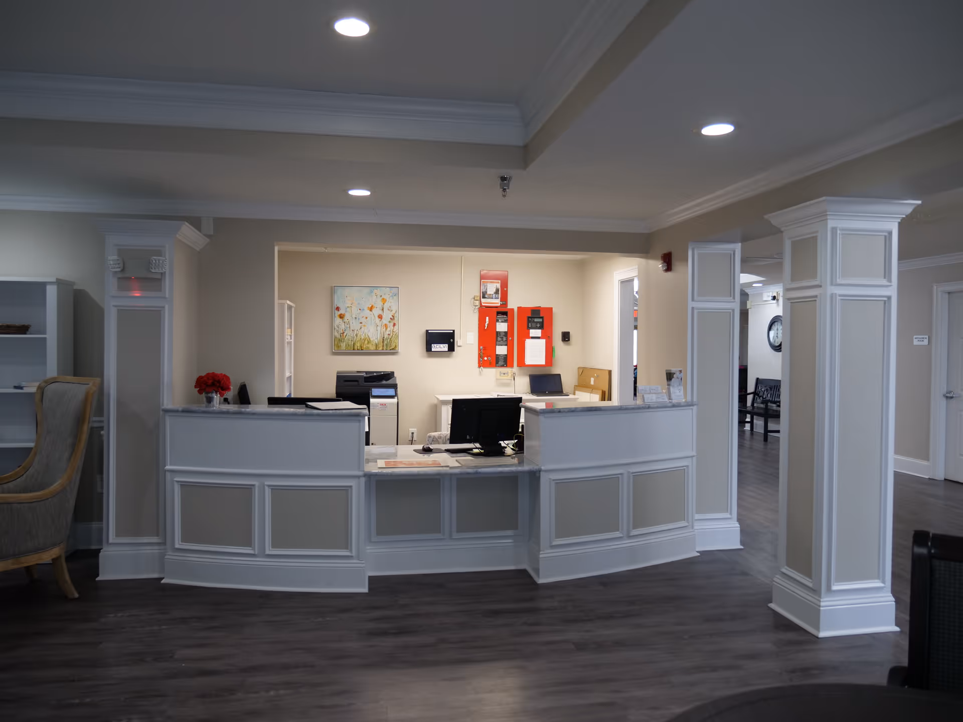 Reception desk area in a senior living facility with a curved white counter, computer monitors, and office equipment. The room has beige walls, white trim, and dark wood flooring. There are decorative columns and a painting of flowers on the wall behind the desk.
