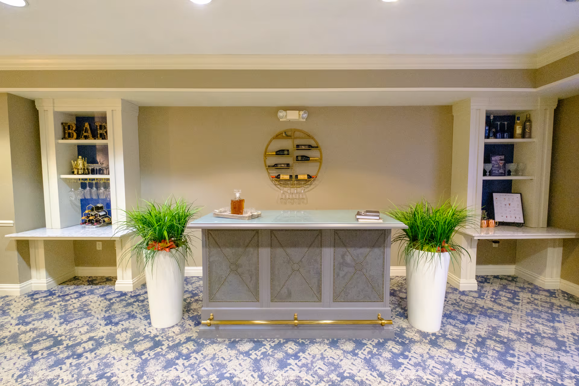 A stylish indoor bar area with a central gray bar counter flanked by two tall white planters with green plants. Behind the bar, there are built-in shelves on either side displaying bottles, glasses, and decorative items. A circular wine rack with several bottles is mounted on the beige wall above the bar. The floor is covered with a blue and white patterned carpet.