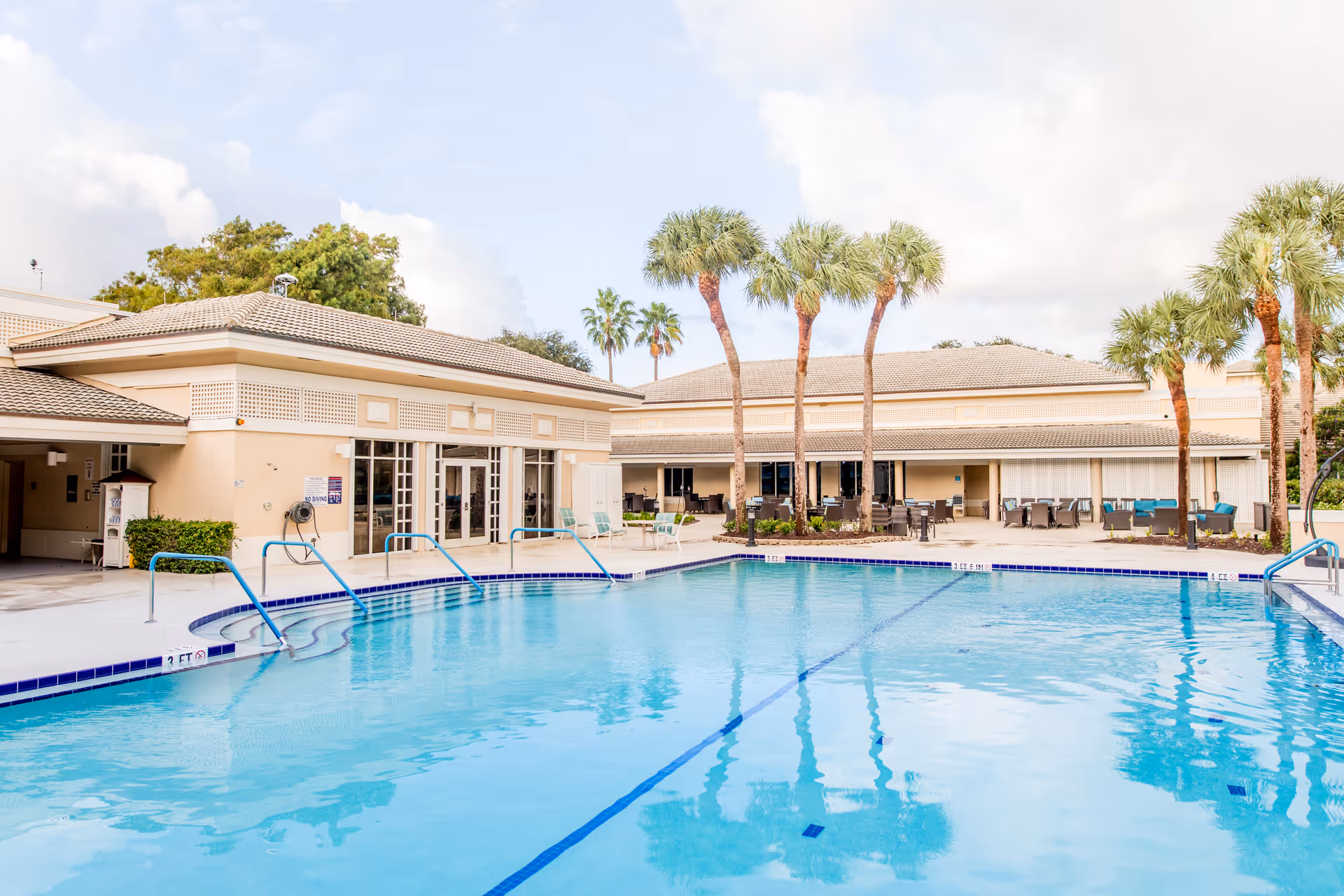 Outdoor swimming pool with clear blue water surrounded by a patio area with chairs and tables. Palm trees are planted around the pool area, and beige buildings with tiled roofs are visible in the background under a partly cloudy sky.