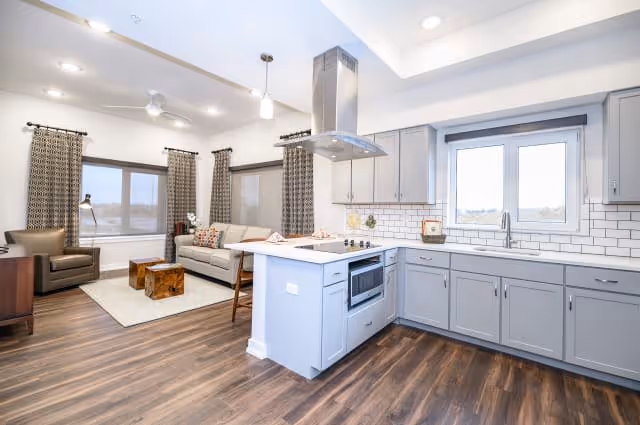 Bright and modern open-concept living area and kitchen with light gray cabinetry, white subway tile backsplash, stainless steel appliances, and wood flooring. The living area features a beige sofa, patterned curtains, a wooden coffee table, an armchair, and large windows allowing natural light.