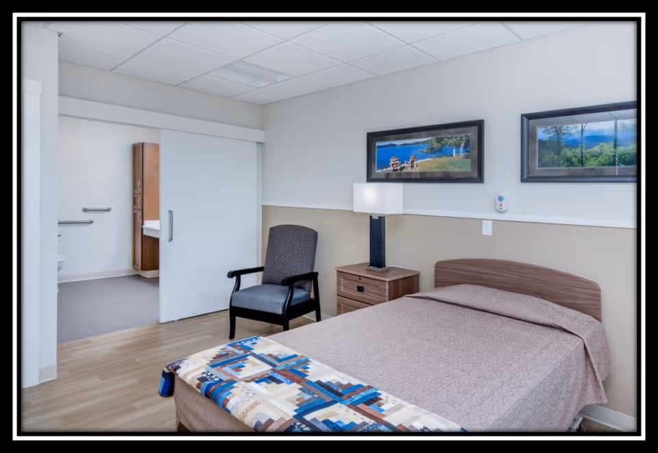 Furnished bedroom with a single bed, nightstand and lamp, an armchair, framed wall art, and an open sliding door revealing an adjacent bathroom.