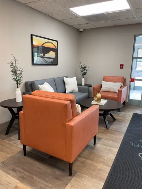 A cozy seating area in a senior living facility with two orange armchairs and a gray sofa arranged around two round black coffee tables. The tables hold decorative items including a gold sculpture and a book. There are two side tables with white vases containing green plants. A framed picture of a bridge at sunset hangs on the beige wall. The floor is wood-patterned, and a glass door is visible in the background.