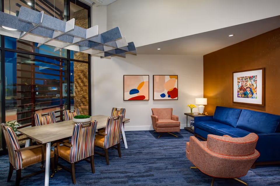 A modern and cozy interior room featuring a wooden dining table with six striped chairs on the left side, and a seating area on the right with a blue sofa, two orange armchairs, a side table with a lamp and flowers, and colorful abstract artwork on the walls.