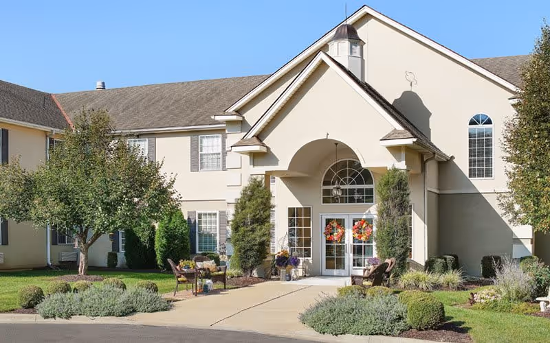 Exterior front view of a senior living facility building with a peaked roof and an arched entrance. The entrance has double glass doors decorated with wreaths. There are trees, shrubs, and landscaped greenery around the building, with a paved walkway leading to the entrance. Outdoor seating is visible near the entrance.