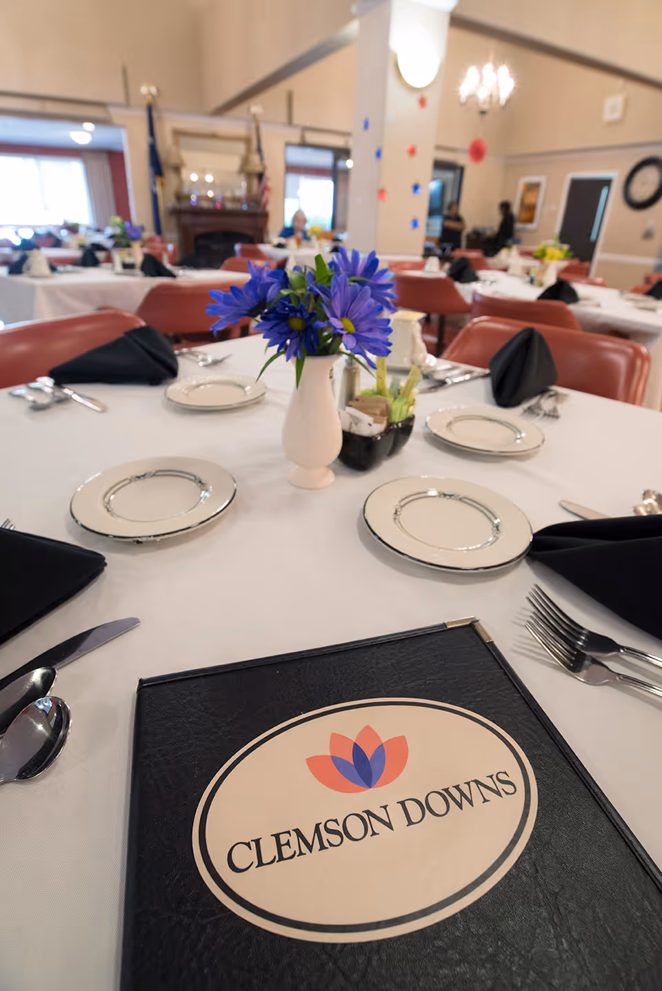 A dining room table set for a meal with plates, silverware, black napkins, a vase of purple flowers, and a Clemson Downs menu in the foreground.