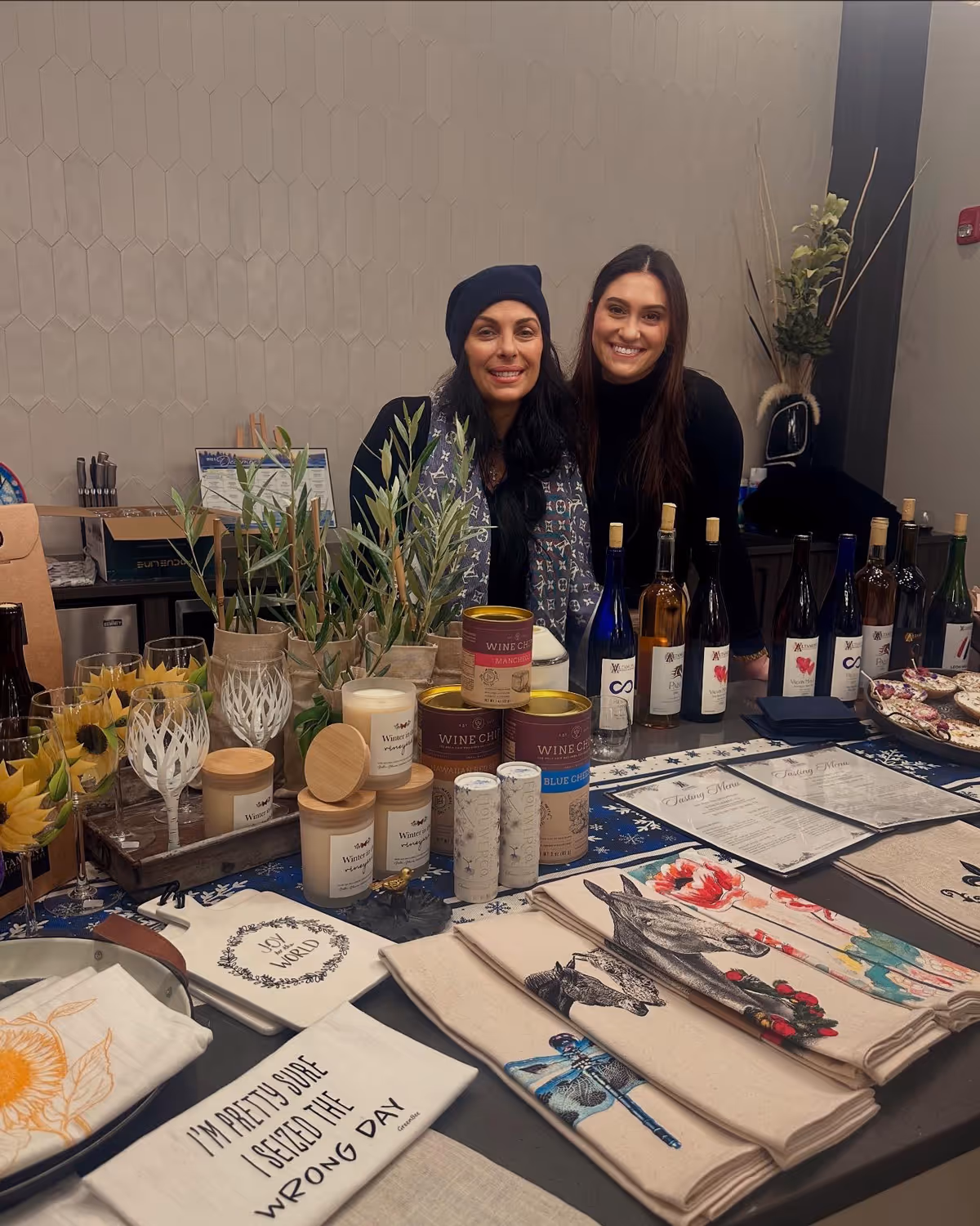 Two women standing behind a table displaying various items including wine bottles, candles, potted plants, decorated cloths, and glasses filled with a yellow dessert. The setting appears to be indoors with a patterned wall in the background.