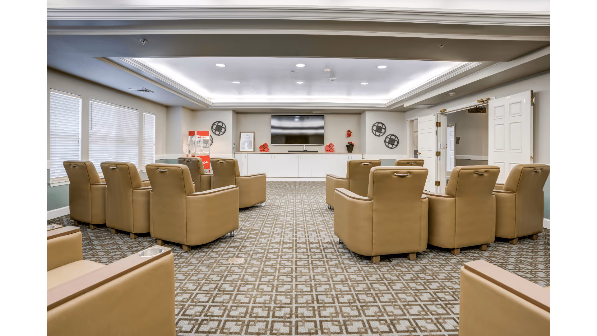 A spacious lounge area with two rows of tan leather armchairs facing a large flat-screen TV mounted on a white cabinet. The room has patterned carpet flooring, a popcorn machine in the corner, and decorative wall art. Large windows with blinds are on the left side, and double doors are open on the right side.