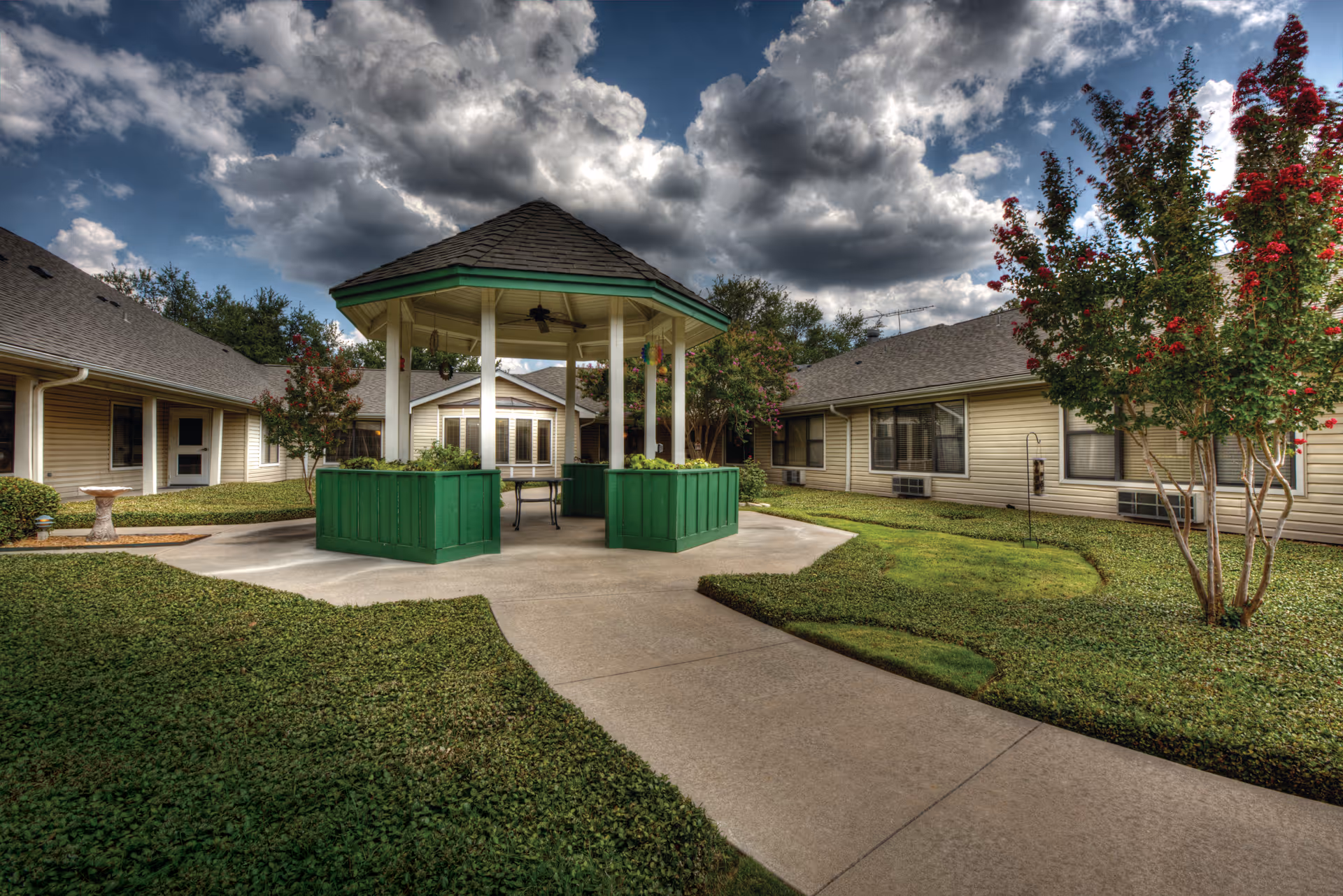 Outdoor courtyard area of a senior living facility with a central green gazebo surrounded by well-maintained grass, trees with red flowers, and beige single-story buildings under a cloudy sky.