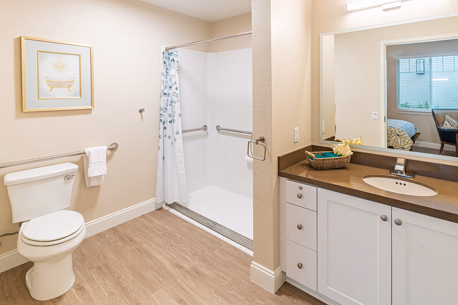 A clean and well-lit bathroom with a white toilet, a walk-in shower with a white curtain featuring a blue and green leaf pattern, and a vanity with a brown countertop and white cabinets. A framed picture of a bathtub hangs on the wall above the toilet. A large mirror above the vanity reflects part of a bedroom with a window and a chair.