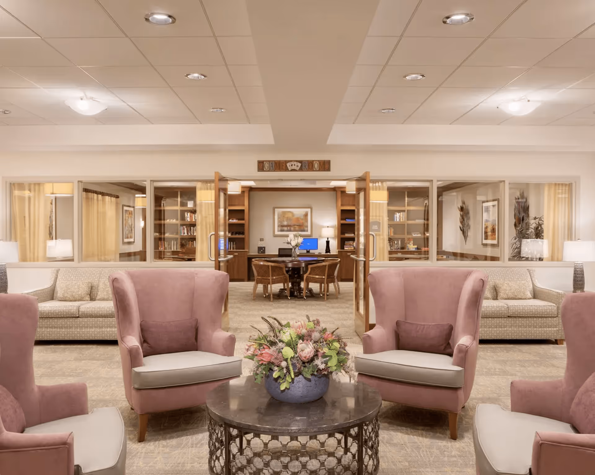 A cozy common area with four pink armchairs arranged around a round coffee table with a floral centerpiece. Behind the seating area, there are two beige sofas with lamps on side tables. In the background, double glass doors lead to a room labeled 'Game Room' with a table, chairs, bookshelves, and a computer.