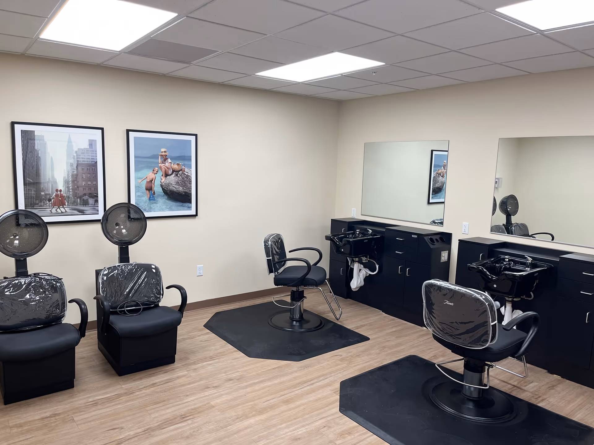 Small salon room with styling chairs, hooded hair dryers, wash sinks, large mirrors and framed artwork on the walls.
