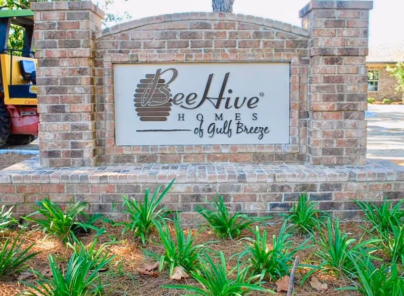 A brick sign for BeeHive Homes of Gulf Breeze, surrounded by green plants and pine straw, with part of a building and a vehicle visible in the background.