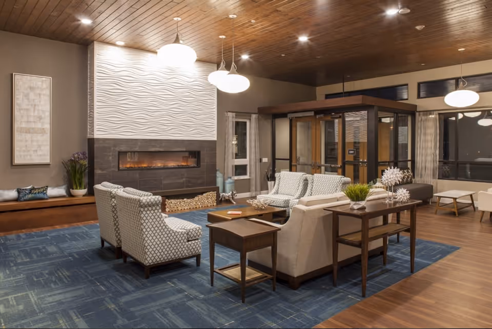 A cozy living room area with patterned armchairs and a sofa arranged around a wooden coffee table on a blue rug. The room features a modern electric fireplace with a textured white wall above it, wooden ceiling with recessed lighting, and large windows with curtains. There is a wooden console table with decorative items and plants behind the sofa. The entrance door with glass panels is visible in the background.