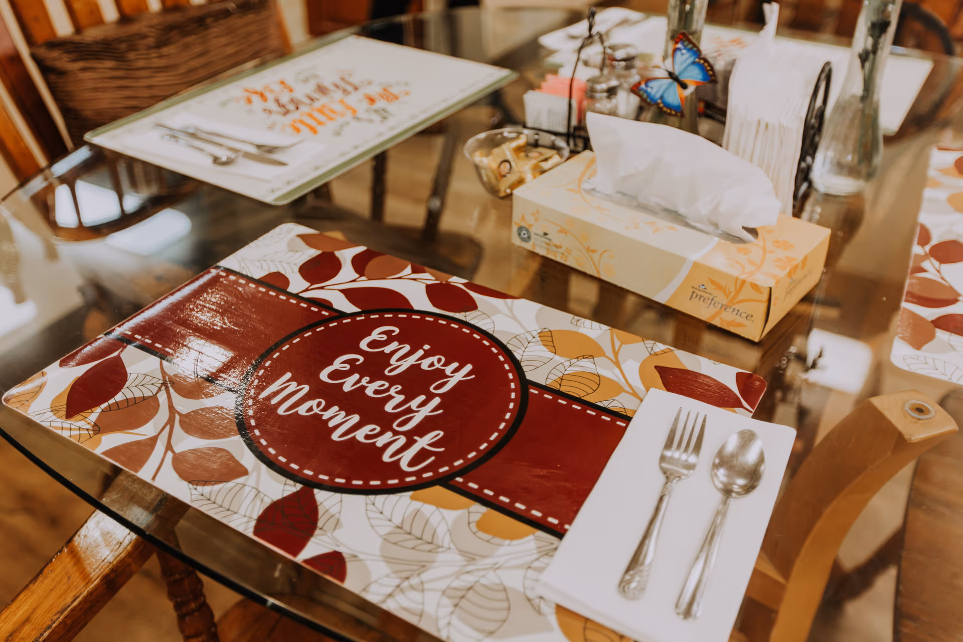 A glass dining table set with placemats featuring autumn leaves and the phrase 'Enjoy Every Moment'. On the table are a fork and spoon on a white napkin, a tissue box, a small bowl with sugar packets, salt and pepper shakers, and a decorative butterfly clip.