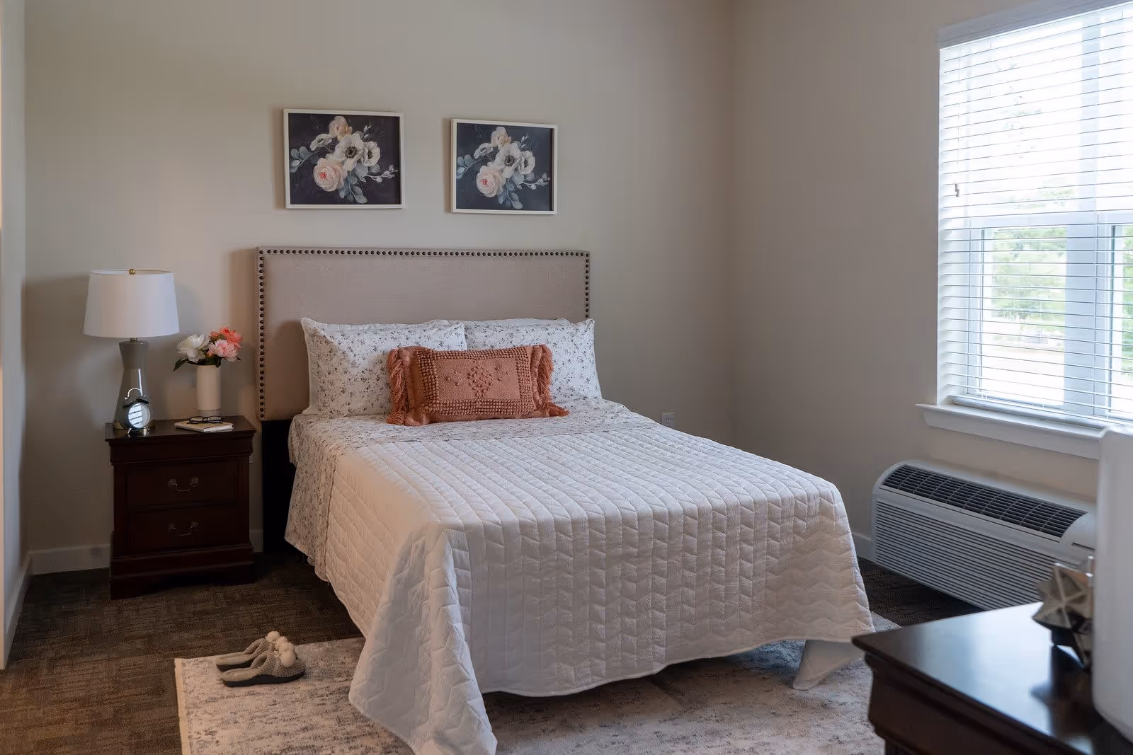 A neatly made bed with a white quilt and floral pillows in a bedroom. There is a wooden nightstand with a lamp, a clock, a book, and a vase with flowers next to the bed. Two framed floral paintings hang on the wall above the headboard. A window with white blinds allows natural light into the room, and slippers are placed on a rug beside the bed.