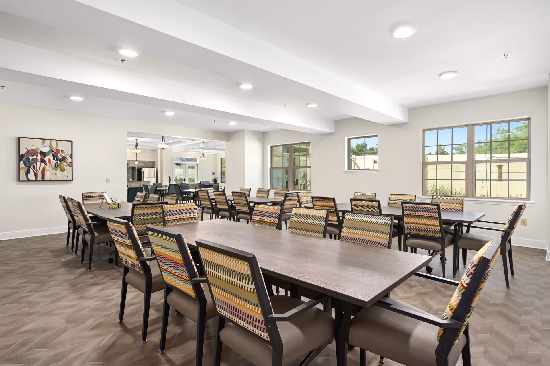 A bright and spacious dining room with multiple rectangular tables and colorful patterned chairs arranged neatly. Large windows allow natural light to fill the room, and a piece of abstract artwork hangs on the wall. The room has a clean, modern design with light-colored walls and wood-patterned flooring.