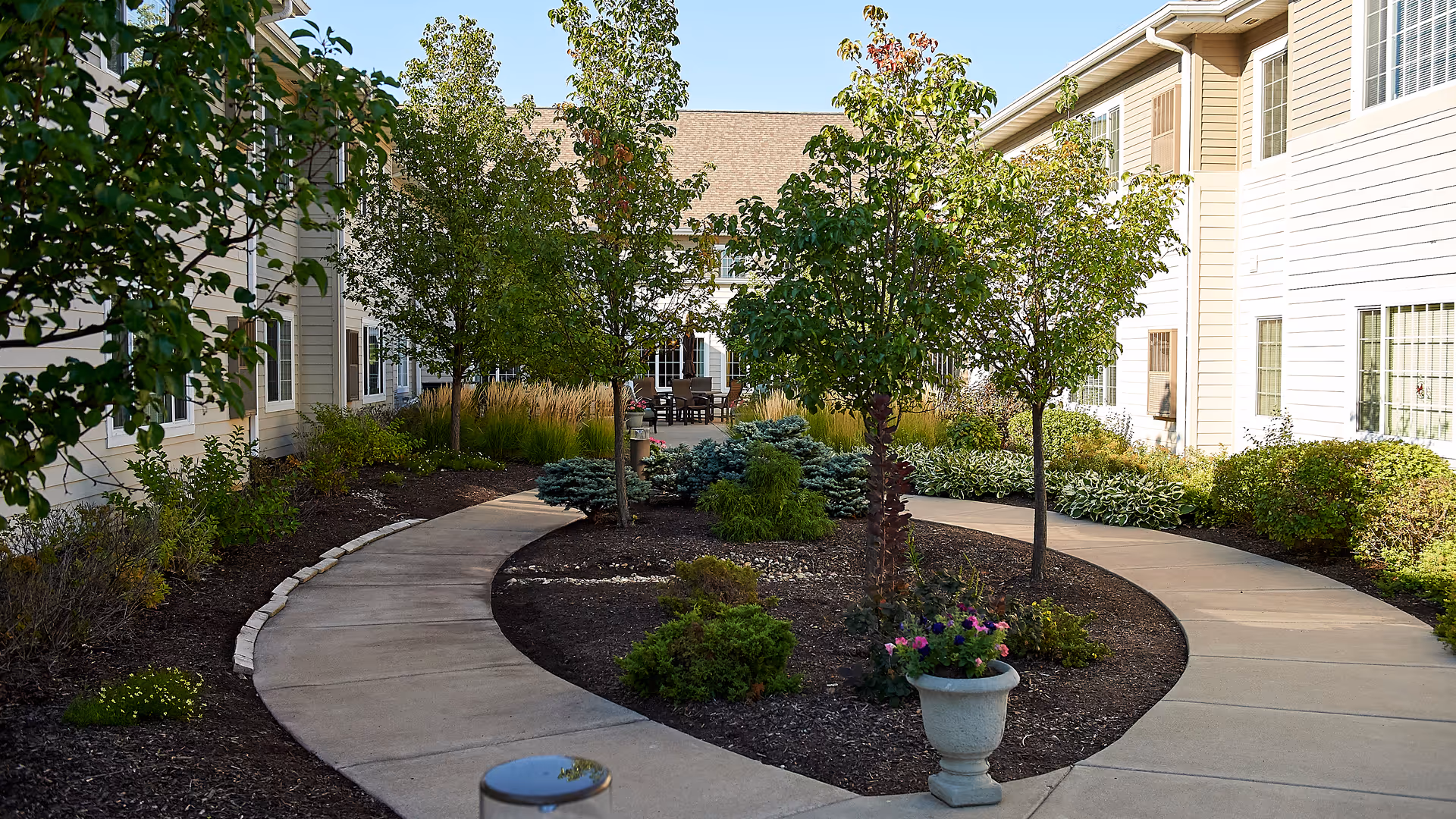 A landscaped courtyard with curved walkways, trees, planters, and outdoor seating between two wings of a senior living building.