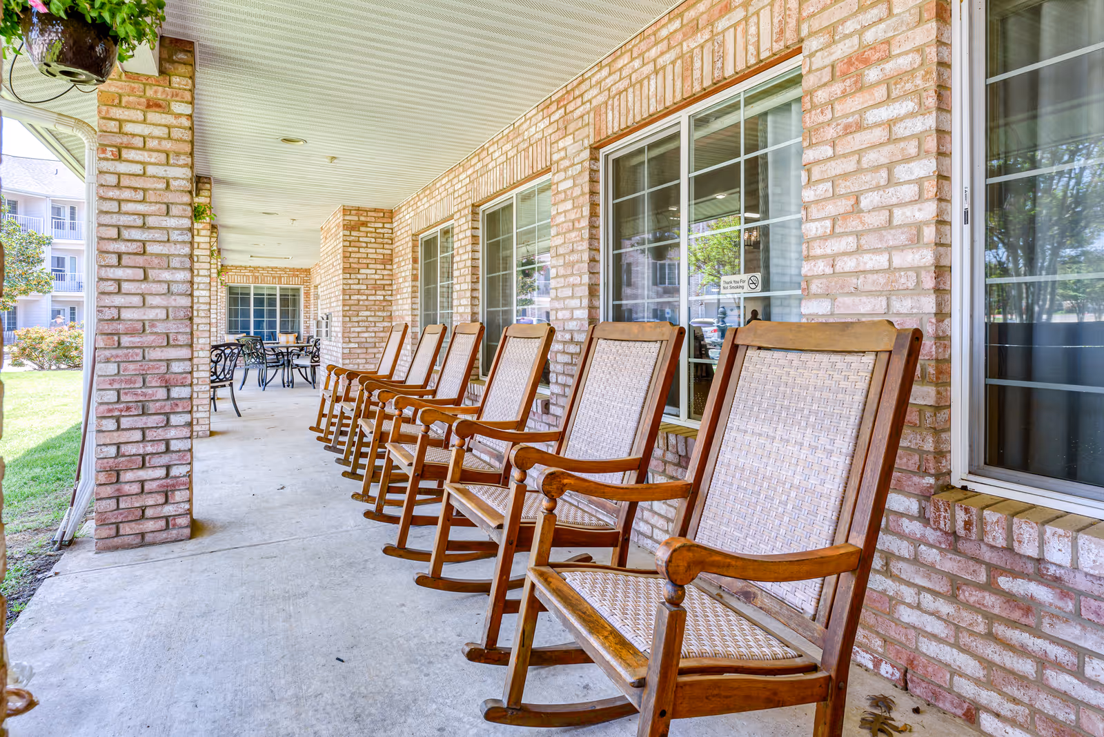 A covered brick patio at a senior living facility with a row of wooden rocking chairs and a table set in the background.