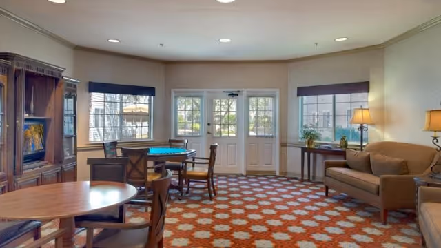 A well-lit common area in a senior living facility featuring a patterned carpet, round wooden tables with chairs, a wooden entertainment center with a TV, a beige sofa, a side table with a lamp, and large windows and glass doors letting in natural light.