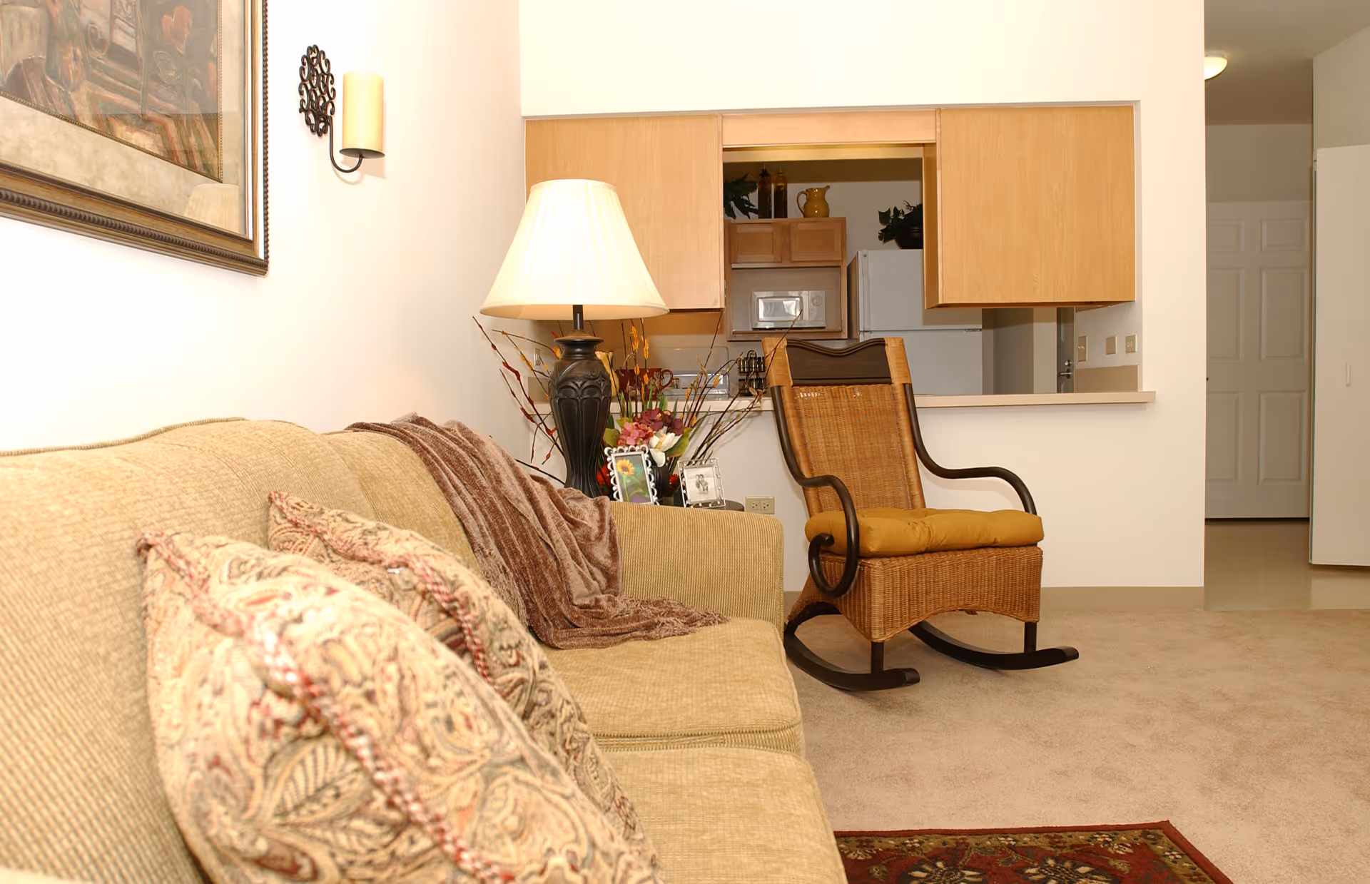 A cozy living room area with a beige sofa adorned with patterned cushions and a brown throw blanket. Next to the sofa is a black table lamp with a cream-colored shade and a floral arrangement. A wooden rocking chair with a yellow cushion is positioned near a pass-through window that opens to a kitchen with wooden cabinets, a microwave, and a refrigerator. The room has beige carpeting and a decorative rug partially visible in the foreground.