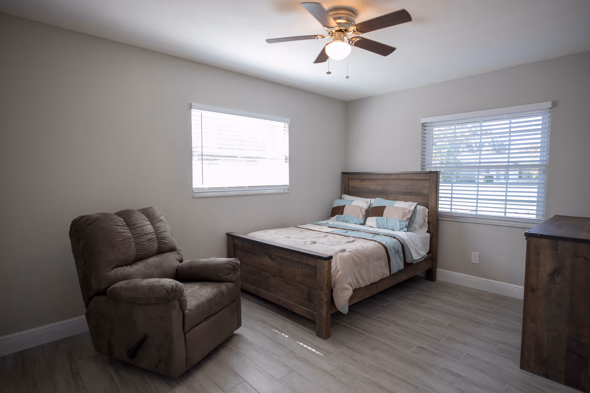 A simple bedroom with a wooden bed frame, a bed made with pillows and a comforter, a brown recliner chair, two windows with blinds, a ceiling fan with light, and a wooden dresser.