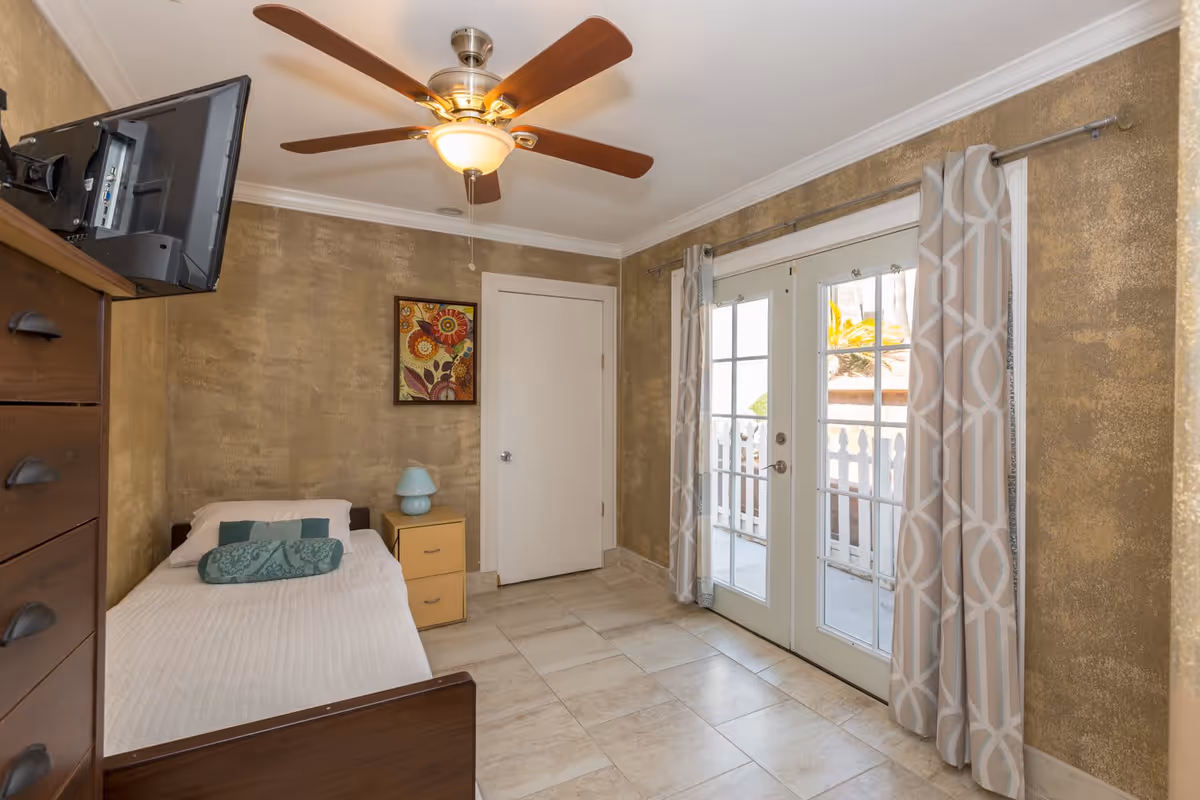 A small bedroom with a single bed covered in a white quilt and a teal bolster pillow. Next to the bed is a light wood nightstand with a blue lamp. A ceiling fan with wooden blades and a light is mounted on the ceiling. There is a wall-mounted TV on the left side above a dark wood dresser. The walls have a textured golden finish, and a framed floral artwork hangs above the nightstand. French doors with patterned curtains lead to an outdoor area with a white picket fence visible outside.