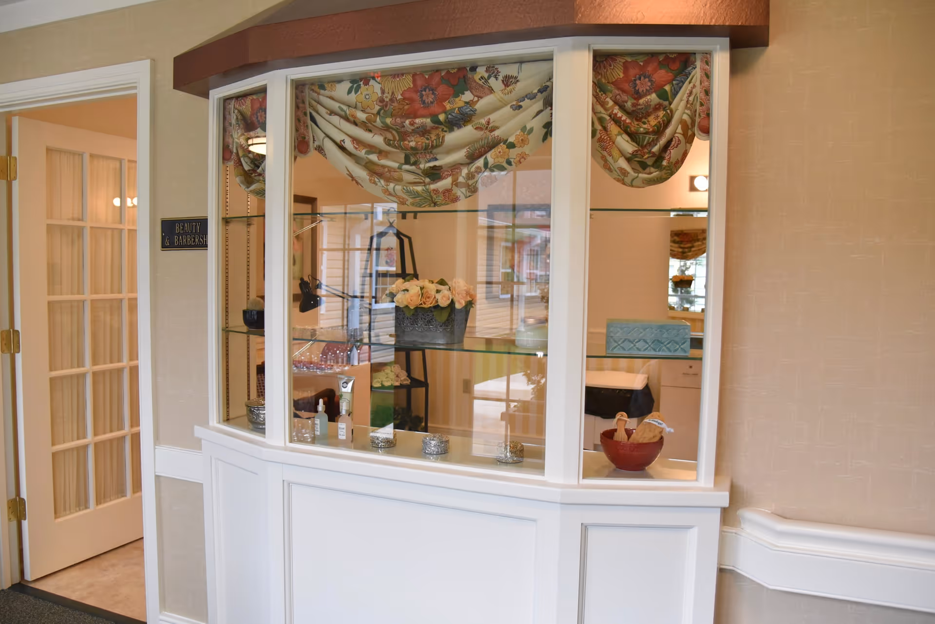Interior view of a beauty and barbershop area in a senior living facility, featuring a glass display case with decorative items such as flowers and small containers. A door with glass panels is open to the left, and the walls are painted beige with white trim.