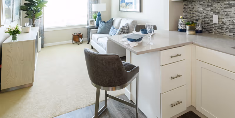 Bright open-plan living area with a sofa, TV console, and a kitchen peninsula with a bar stool.