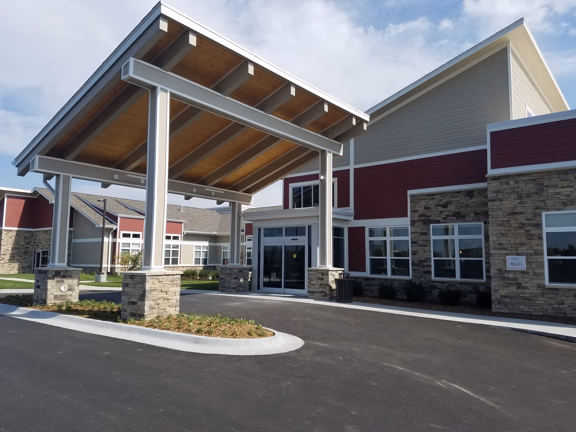Entrance of a senior living facility with a covered drop-off area supported by stone pillars. The building has a combination of stone and red siding with multiple windows and a clear sky in the background.