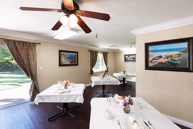 A bright dining room with four tables covered in white tablecloths, each set with plates, glasses, silverware, and small floral centerpieces. The room has beige walls adorned with framed paintings of boats and coastal scenes. Large windows with patterned curtains let in natural light, and a ceiling fan with lights is mounted above. The floor is dark wood, and a door opens to a green outdoor area.