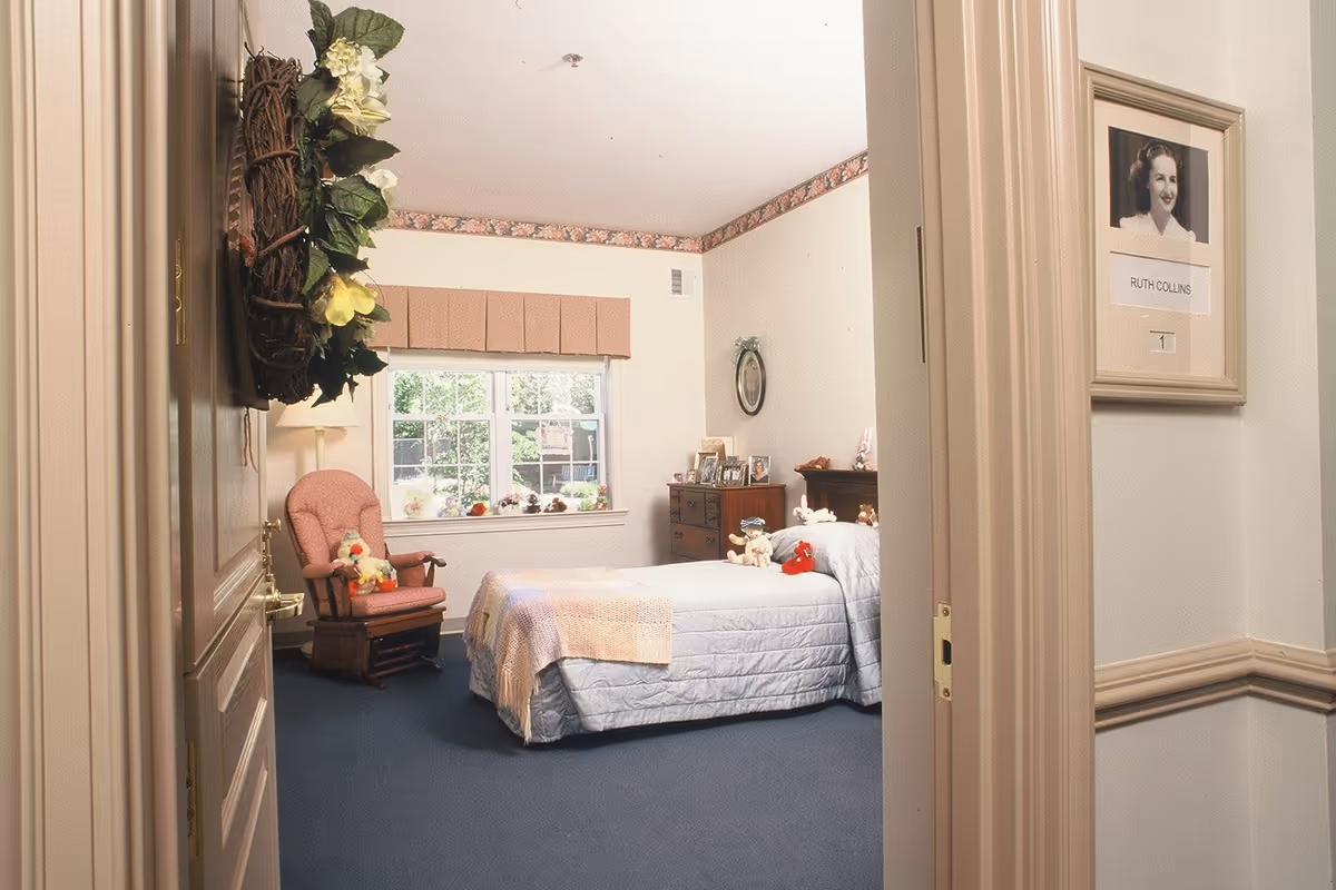 View into a cozy bedroom with a single bed covered in a light blue quilt and a pastel blanket. Several stuffed animals are placed on the bed. A wooden dresser with framed photos and a small mirror hangs on the wall. A pink cushioned rocking chair with a stuffed duck sits near a window with a valance. The room has floral wallpaper border near the ceiling and blue carpeted floor. A framed photo and nameplate reading 'Ruth Collins' is mounted on the wall outside the room.